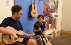 young girl with down syndrome holds a guitar while sitting next to her brother who is playing a guitar